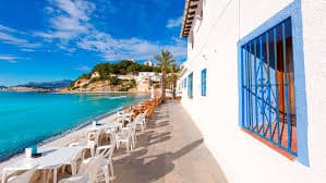 A sunny outdoor café terrace with white tables and chairs lined up along a white building with a bright blue-barred window, overlooking the turquoise ocean and a green cliff. This aspirational image, used by a crypto learning platform, symbolizes the successful lifestyle and financial freedom achieved after mastering decentralized finance and focusing on crypto learning and investment.